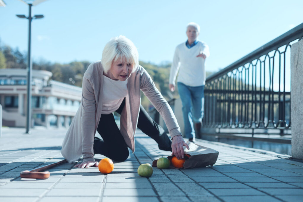 Older adult female fell outdoors. Dropping a paper bag and some fruit rolling out of it. Male older adult running up behind her to help. Fall risk assessments can help to reduce the risk of falls. 