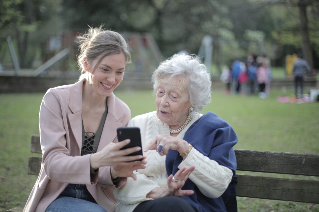 Physician and older adult sitting on a park bench looking at a smartphone. They are both feeling happy and proud, likely because their fall prevention efforts have been successful.  The background is blurred out. 