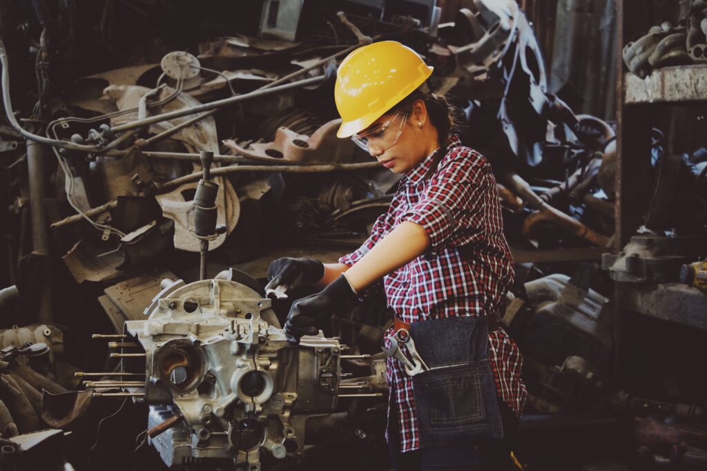 Female factory worker, working on an engine. She is wearing her hard hat, safety glasses, and gloves. She is awaiting the results from her occupational testing. 